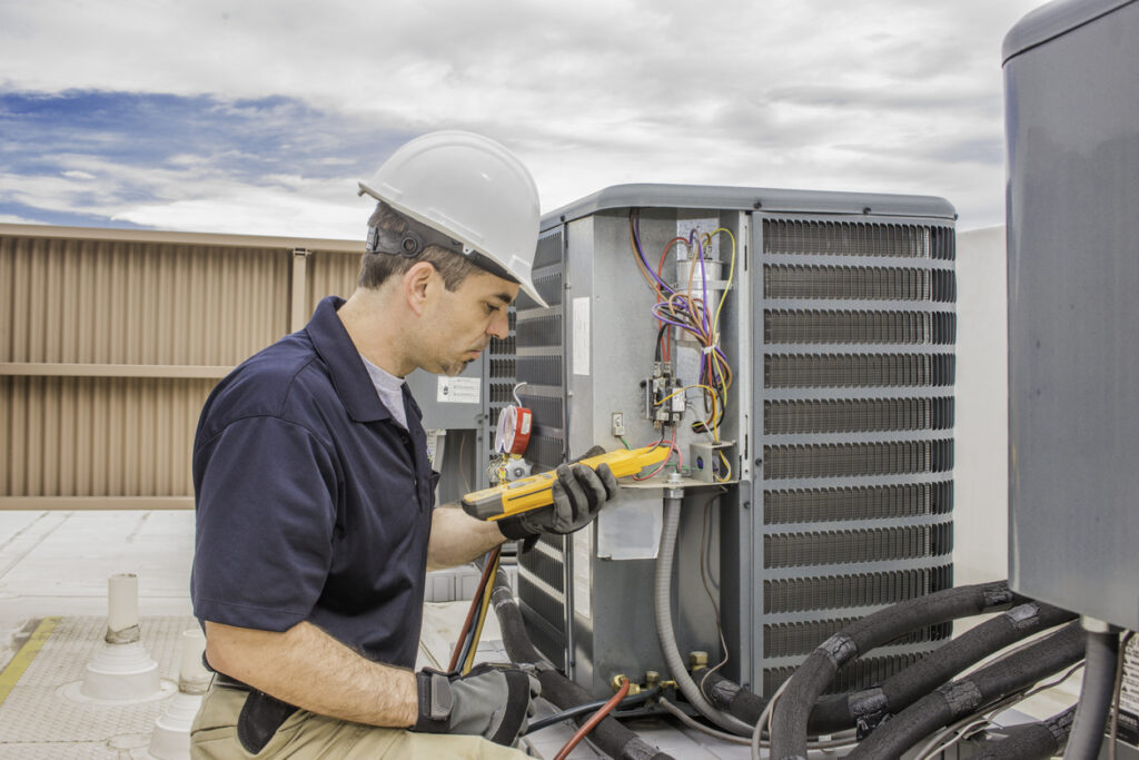 Trained HVAC technician holding a voltage meter, performing preventative maintenance on a air conditioning condenser unit.