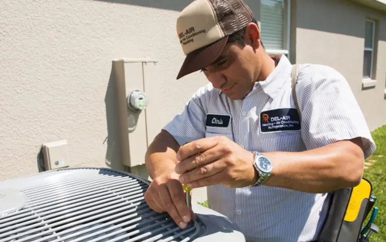 Del-Air AC technician repairing an air conditioner next to a white stucco home.