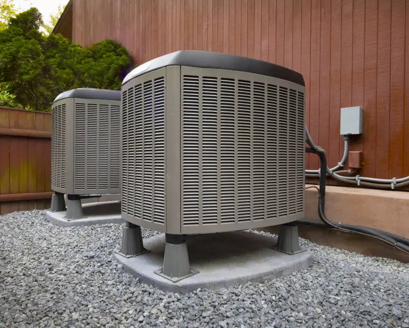 Two outdoor HVAC units installed next to a brown home with vertical siding.