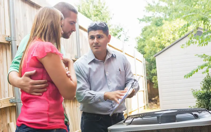 Technician speaking with two homeowners while standing in front of an outdoor HVAC unit. Wooden fence behind them.