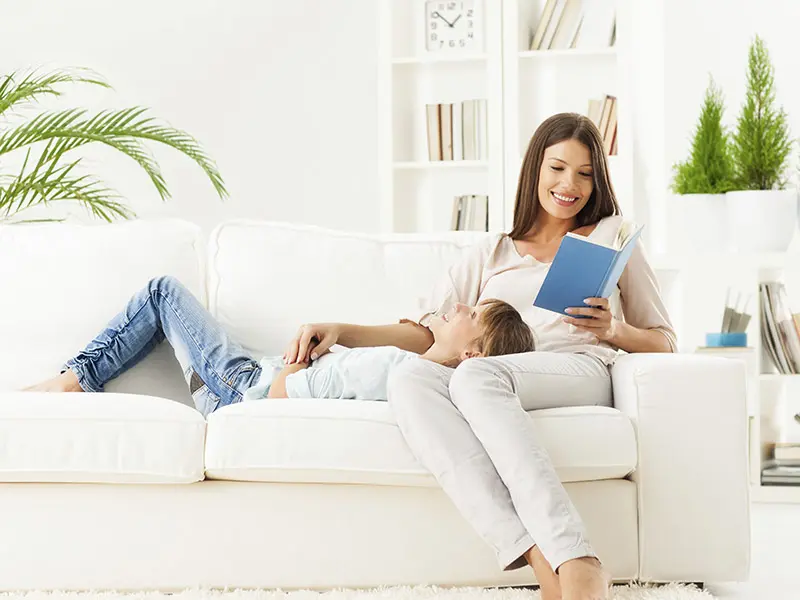 mom and daughter comfortable on a white couch with white wall and bookcase and houseplants in background.
